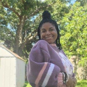 May include: A woman wearing a purple and white striped jumper with a crocheted texture. She is wearing blue jeans and has her hair pulled back in a bun.