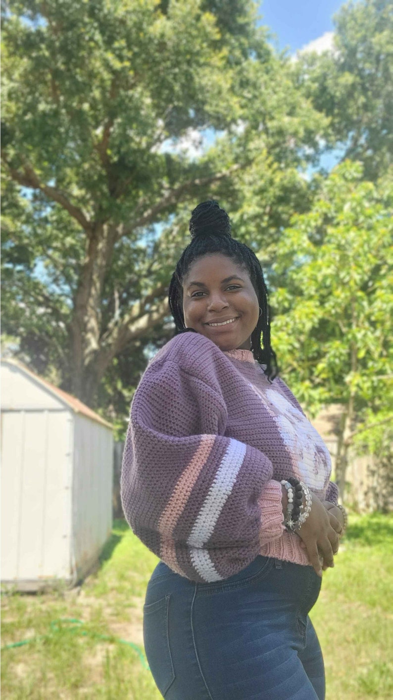 May include: A woman wearing a purple and white striped jumper with a crocheted texture. She is wearing blue jeans and has her hair pulled back in a bun.
