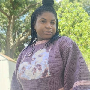 May include: A woman wearing a purple crocheted sweater with a white and pink design. The sweater has long sleeves with a white and pink stripe at the cuffs. The woman is wearing blue jeans.