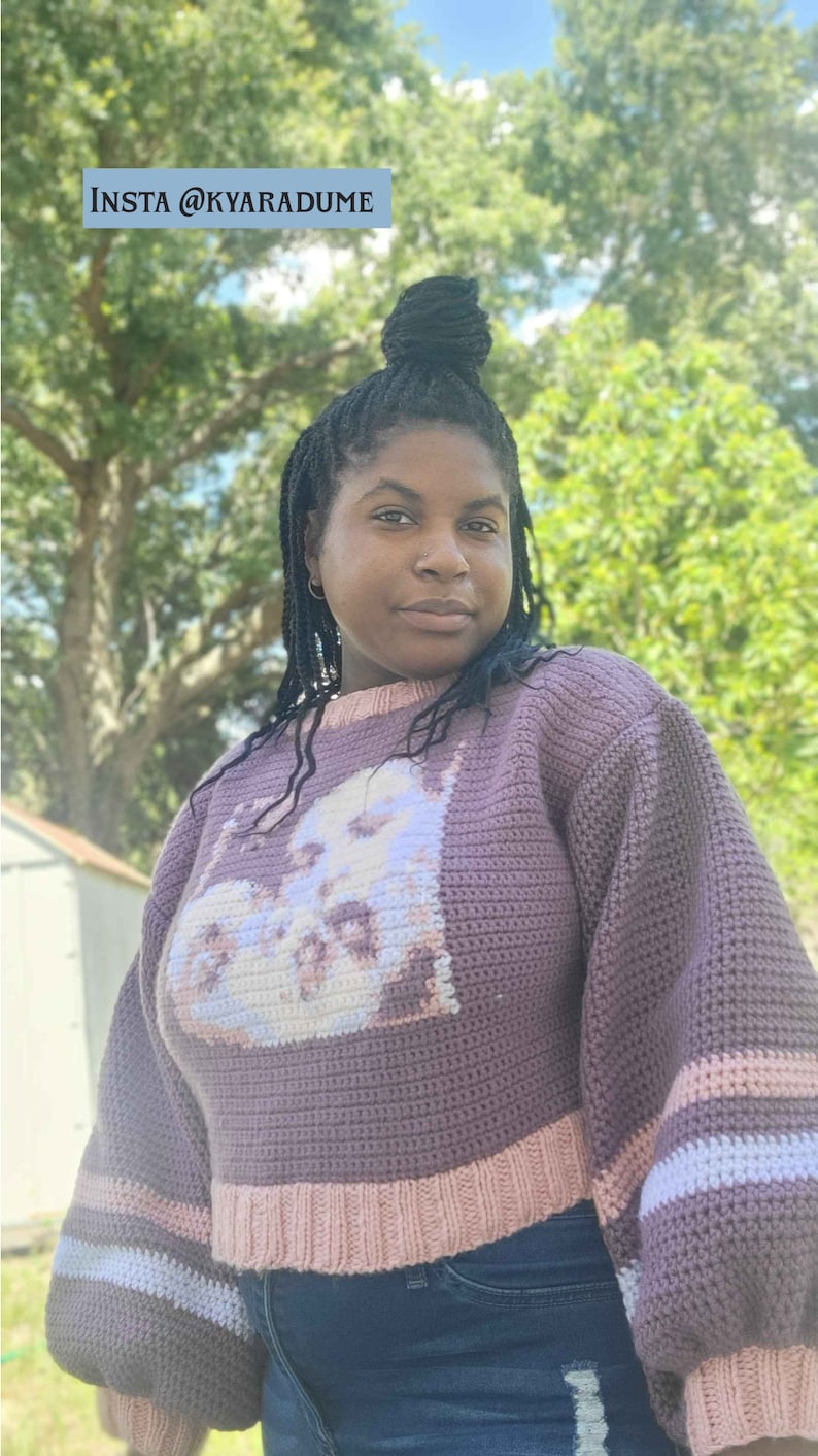 May include: A woman wearing a purple crocheted sweater with a white and pink design. The sweater has long sleeves with a white and pink stripe at the cuffs. The woman is wearing blue jeans.