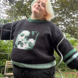 May include: A person wearing a black and green jumper with a green and white graphic design. The jumper has a green and white stripe on the sleeve. The person is wearing black trousers and is standing in front of a green background. The text "TikTok @LEIGHTONS.ARTSPACE" is visible at the bottom of the image.