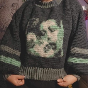 May include: A dark green and light green crocheted jumper with a graphic image of a face.