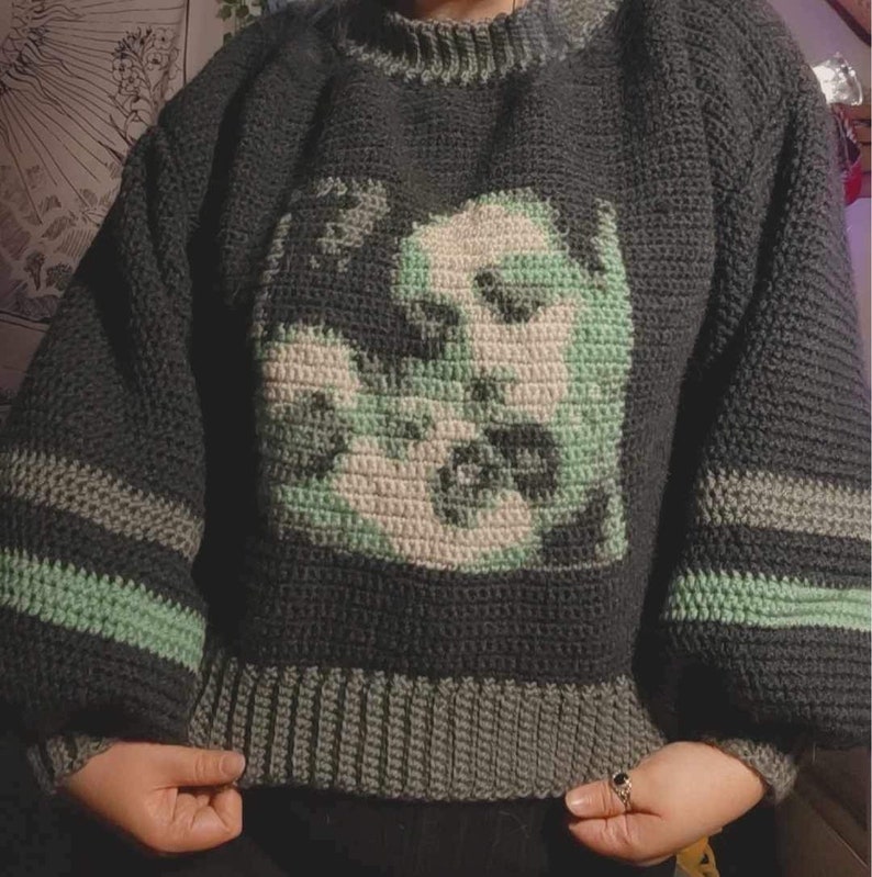May include: A dark green and light green crocheted jumper with a graphic image of a face.