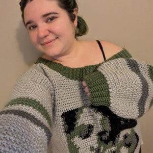 May include: A person wearing a grey and green crocheted sweater with a black and white graphic of a creature's face. The sweater has a wide, off-the-shoulder neckline and long sleeves. The text "TikTok @MOSSY_CRYPTID" is visible in the top right corner of the image.