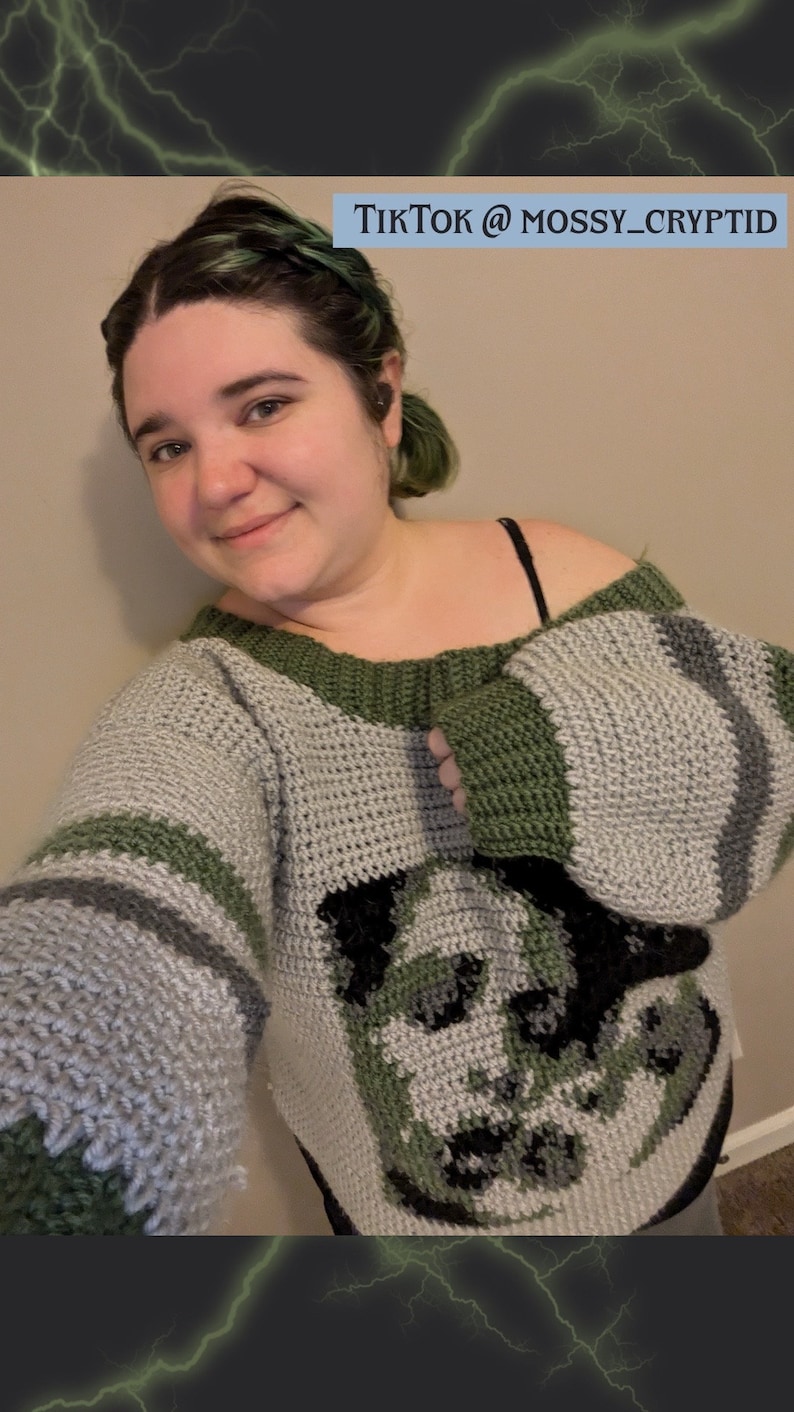 May include: A person wearing a grey and green crocheted sweater with a black and white graphic of a creature's face. The sweater has a wide, off-the-shoulder neckline and long sleeves. The text "TikTok @MOSSY_CRYPTID" is visible in the top right corner of the image.