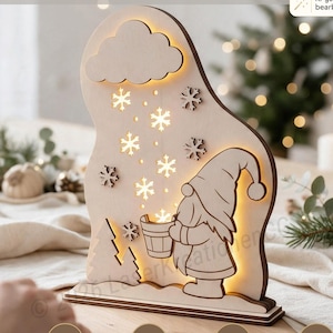 Può includere: Un pezzo decorativo in legno chiaro tagliato al laser raffigurante uno gnomo che tiene un secchio sotto una nuvola con fiocchi di neve che cadono. Il design è illuminato da dietro, emettendo una luce calda. Il testo in alto recita LASER CUT FILE by Laser Kreationen CG.