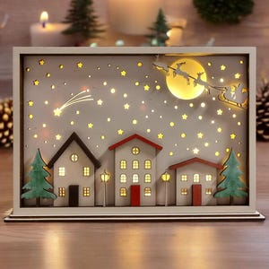 Könnte beinhalten: Eine weiße Holz-Weihnachtsdorf-Szene mit einem beleuchteten Mond und Sternen. Die Szene zeigt drei Häuser mit roten Dächern und Fenstern, einen Sternschnuppen und einen Schlitten mit Rentieren, die am Himmel fliegen.