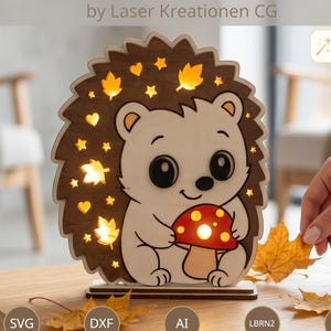 Può includere: Un file di taglio laser raffigurante un riccio in legno illuminato che tiene un fungo rosso con macchie bianche. Il riccio è circondato da stelle e foglie ritagliate. Il testo include LASER CUT FILE, by Laser Kreationen CG, e i formati SVG, DXF, AI, LBRN2, PDF.
