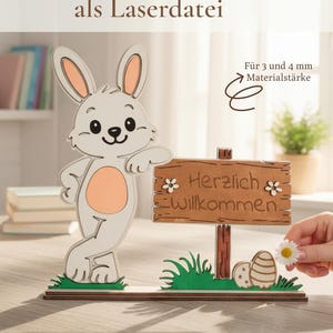 Könnte beinhalten: Osterhase Dekoration aus Holz mit einem Willkommensschild. Der Hase ist weiß mit einem pfirsichfarbenen Bauch und Ohren. Das Schild trägt die Aufschrift "Herzlich Willkommen" mit Blumen. Die Basis umfasst Gras und Ostereier.