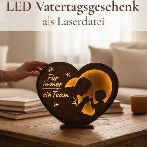 Op de afbeelding: Een hartvormige houten LED-lamp met een silhouet van een ouder en kind. De lamp heeft de tekst "Für immer ein Team" en "LED Vatertagsgeschenk als Laserdatei". De lamp staat op een houten tafel.