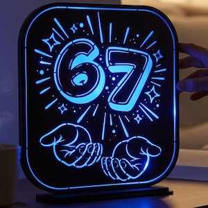 May include: A black, rectangular LED light with the number "67" illuminated in blue. The light has a stylized design with hands and starbursts. It sits on a wooden nightstand next to a cup and an open book.