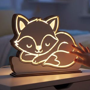 Könnte beinhalten: Ein hölzernes Nachtlicht in Fuchsform, das ein warmes Licht ausstrahlt. Das Licht steht auf einem weißen Nachttisch. Der Fuchs liegt mit geschlossenen Augen. Das Licht ist an eine Steckdose angeschlossen.
