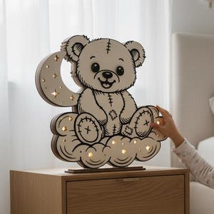 Op de afbeelding: Een houten nachtlampje in de vorm van een teddybeer op een wolk met een maan. Het licht staat op een houten nachtkastje. De beer heeft gestikte details en is omgeven door kleine lampjes. Het kleurenschema is neutraal met warme verlichting.