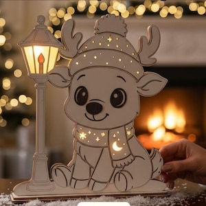 Könnte beinhalten: Eine lasergeschnittene Holznachtlampe mit einem Cartoon-Rentier, das eine Wintermütze und einen Schal trägt und neben einer Straßenlaterne steht. Das Design enthält Stern- und Mondausschnitte. Text: Laser Cut File - Night Lamp - Multi-Layer Design.