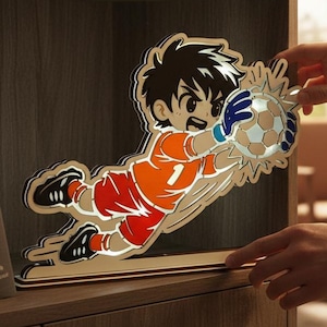 Puede incluir: Un recorte de madera de un portero de fútbol de dibujos animados con una camiseta naranja y pantalones cortos rojos, lanzándose a atrapar un balón de fútbol. El portero tiene el pelo negro y guantes azules. El recorte está sobre una base de madera.