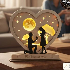 Può includere: Un file per taglio laser a forma di cuore con la silhouette di una persona inginocchiata per fare una proposta di matrimonio sotto una luna e stelle. La base include il testo 'Für immer wir' e due piccole icone a forma di cuore.