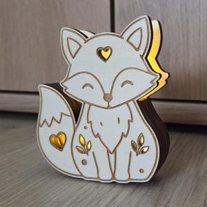 May include: A layered wooden fox-shaped light fixture with a white front panel featuring cutouts of a heart on the forehead, a heart on the tail, and leaf motifs on the body, emitting a warm yellow glow from the interior layers.
