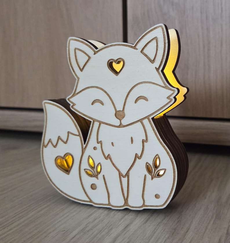 May include: A layered wooden fox-shaped light fixture with a white front panel featuring cutouts of a heart on the forehead, a heart on the tail, and leaf motifs on the body, emitting a warm yellow glow from the interior layers.