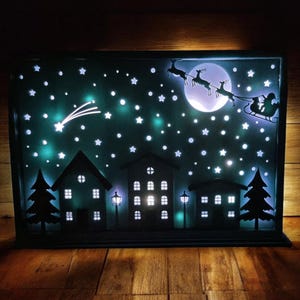 May include: A black metal silhouette of a winter scene with a white moon, stars, and a flying Santa Claus with reindeer. The scene is illuminated with white LED lights.
