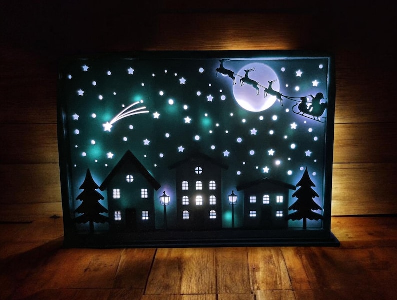 May include: A black metal silhouette of a winter scene with a white moon, stars, and a flying Santa Claus with reindeer. The scene is illuminated with white LED lights.
