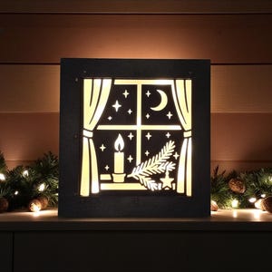 May include: A black, square, illuminated Christmas decoration featuring a window design. The window shows a candle, stars, a crescent moon, and a sprig of pine. The decoration is set against a backdrop of Christmas greenery and lights.