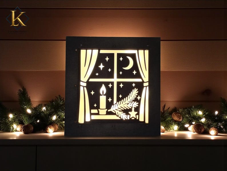 May include: A black, square, illuminated Christmas decoration featuring a window design. The window shows a candle, stars, a crescent moon, and a sprig of pine. The decoration is set against a backdrop of Christmas greenery and lights.