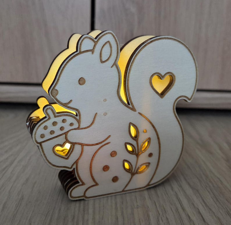 May include: A light-colored wooden squirrel-shaped lamp with a warm internal glow. The squirrel holds an acorn with a heart-shaped cutout, and its tail features a heart cutout and a leaf pattern. The edges are dark, highlighting the silhouette.