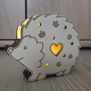 May include: A light-colored wooden hedgehog-shaped lamp with a warm internal glow. The cutout design features a central heart shape, several small circular holes, and maple leaf silhouettes, creating a soft illumination through the wood.