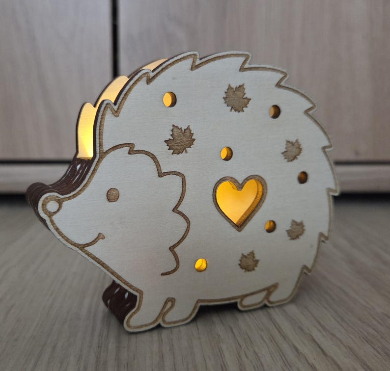 May include: A light-colored wooden hedgehog-shaped lamp with a warm internal glow. The cutout design features a central heart shape, several small circular holes, and maple leaf silhouettes, creating a soft illumination through the wood.
