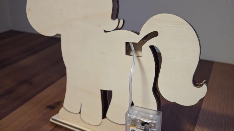 K&ouml;nnte beinhalten: Eine Holz-Pony-f&ouml;rmige Leuchte. Das Pony besteht aus hellfarbenem Holz mit dunkelbraunen Akzenten. Ein klarer Draht erstreckt sich vom Pony zu einer kleinen, durchsichtigen Plastikbox. Das Pony steht auf einem rechteckigen Sockel.