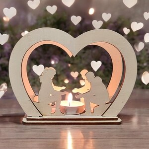 May include: A wooden heart-shaped candle holder with two cutout figures holding hands inside. The candle holder is white and has a small tea light candle in the center.