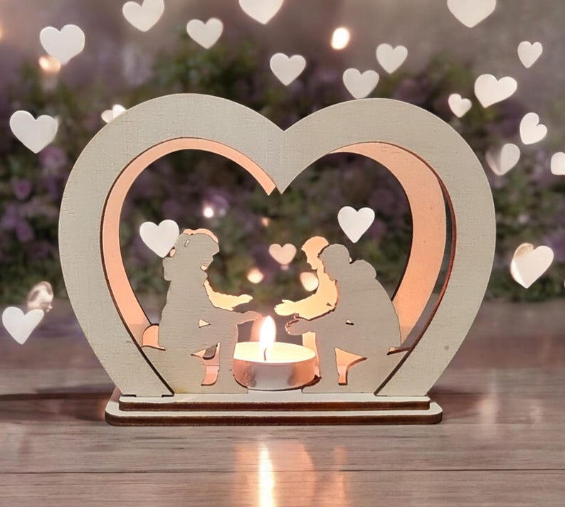 May include: A wooden heart-shaped candle holder with two cutout figures holding hands inside. The candle holder is white and has a small tea light candle in the center.