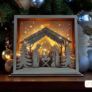 Puede incluir: Una escena de nacimiento de madera cuadrada e iluminada con recortes en capas de la sagrada familia, pinos y un cielo estrellado. La luz cálida emana desde detrás de las siluetas, creando un efecto de profundidad dentro del marco.