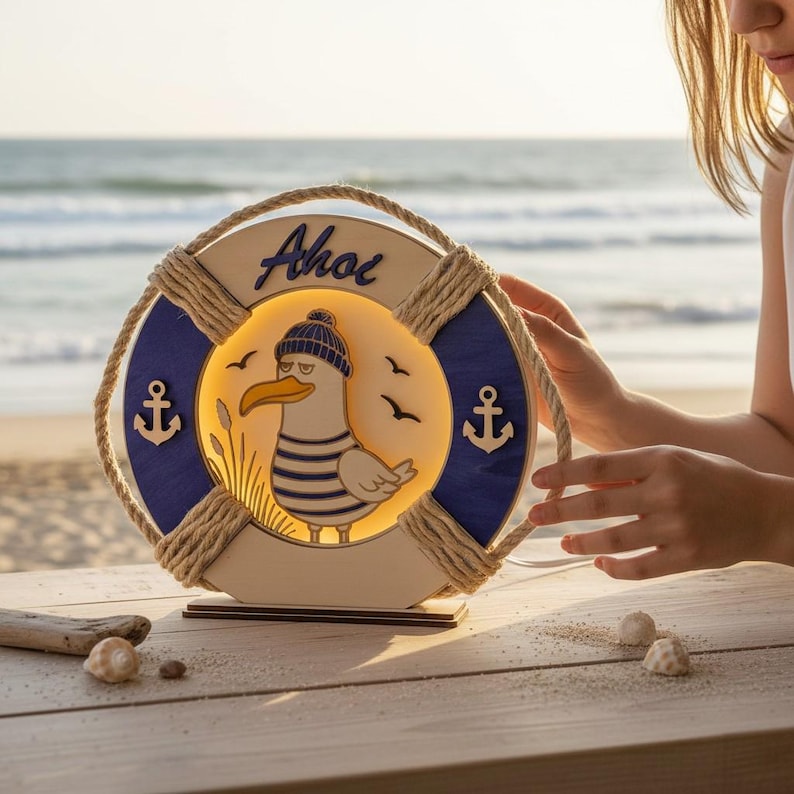 Pu&ograve; includere: Lampada in legno a tema nautico a forma di salvagente, con un'illustrazione di gabbiano e la scritta "Ahoi". La lampada ha uno schema di colori blu e bianco, con dettagli in corda e dettagli di ancora. La lampada &egrave; illuminata dall'interno, creando un bagliore caldo.
