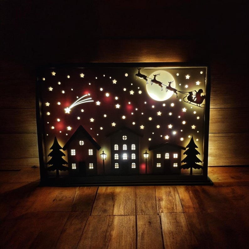 May include: A wooden Christmas scene light-up decoration featuring a silhouette of Santa Claus and reindeer flying over a town with stars and a shooting star in the night sky.