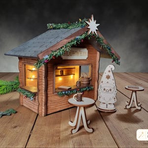 May include: A wooden miniature Gl&uuml;hwein stall featuring a dark roof, illuminated interior, and a sign reading GL&Uuml;HWEIN. The set includes a gnome figure, two small round tables, and miniature mugs. The structure is decorated with green garland.
