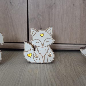 May include: Three light-up wooden animal figures featuring a squirrel holding an acorn, a fox, and a hedgehog. Each white cutout ornament has brown edges and heart-shaped or leaf-shaped openings that emit a warm yellow glow from within.