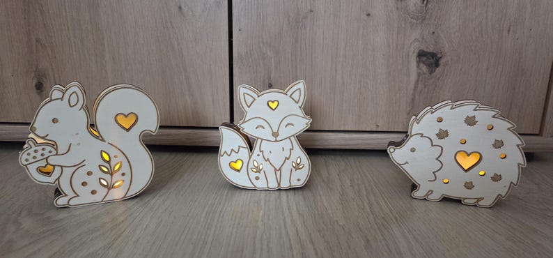 May include: Three light-up wooden animal figures featuring a squirrel holding an acorn, a fox, and a hedgehog. Each white cutout ornament has brown edges and heart-shaped or leaf-shaped openings that emit a warm yellow glow from within.