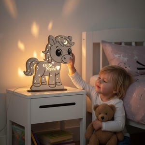 K&ouml;nnte beinhalten: Ein h&ouml;lzernes Nachtlicht in Form eines Pferdes mit Mond- und Sternendesign. Das Licht hat die Worte "Gute Nacht" und ein Herz. Das Licht steht auf einem wei&szlig;en Nachttisch neben einem Kind, das einen Teddyb&auml;ren h&auml;lt.