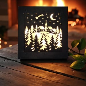 May include: A black, square, illuminated decorative lantern with a winter village scene. The design features a cutout of a village, trees, stars, a crescent moon, and a shooting star. The lantern is placed on a wooden surface.