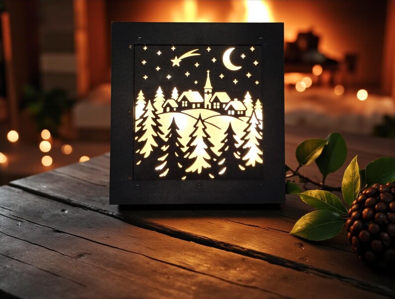 May include: A black, square, illuminated decorative lantern with a winter village scene. The design features a cutout of a village, trees, stars, a crescent moon, and a shooting star. The lantern is placed on a wooden surface.