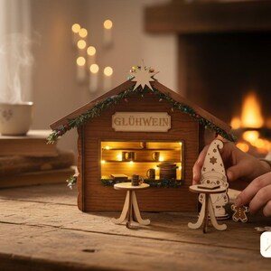 May include: A miniature wooden Gl&uuml;hwein stall featuring a peaked roof with a star, illuminated interior shelves, and two small round tables. The structure is decorated with green garland and includes a small wooden gnome figure and a gingerbread man.