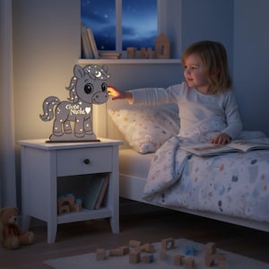 K&ouml;nnte beinhalten: Graues Holz-Nachtlicht in Pferdeform mit den Worten "Gute Nacht" und sternf&ouml;rmigen Ausschnitten. Das Nachtlicht steht auf einem wei&szlig;en Nachttisch. Ein Kind greift nach dem Licht.