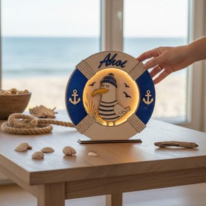 Pu&ograve; includere: Lampada in legno a tema nautico con un gabbiano dei cartoni animati con cappello e camicia a righe, all'interno di una forma di salvagente blu e bianco. La parola "Ahoi" &egrave; scritta in alto. La lampada &egrave; su una base di legno.