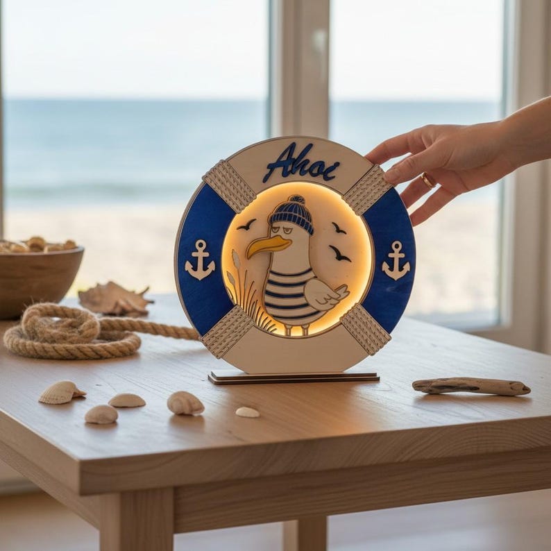 Pu&ograve; includere: Lampada in legno a tema nautico con un gabbiano dei cartoni animati con cappello e camicia a righe, all'interno di una forma di salvagente blu e bianco. La parola "Ahoi" &egrave; scritta in alto. La lampada &egrave; su una base di legno.