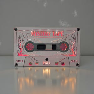 May include: A light-colored wooden cassette tape with the words "MYSTERY TAPE" and "1984" etched into the surface. The design includes red illuminated accents and decorative tree-like patterns. The cassette tape has a Side A and Side B label.