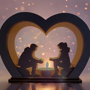 May include: A wooden heart-shaped candle holder with two silhouettes of people sitting inside. The candle holder is lit and the light shines through the silhouettes. The background is a soft purple with white dots.