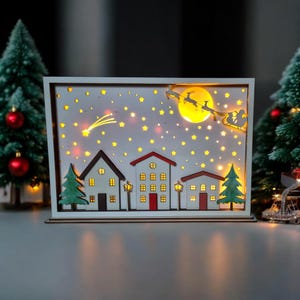 May include: A white wooden Christmas village scene with a lit-up moon, stars, and a shooting star. The scene features three houses with red roofs and windows, a Christmas tree, and a sleigh with Santa and reindeer flying overhead.