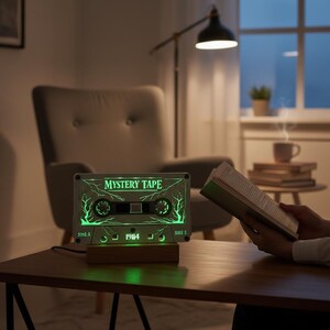 May include: A retro-style cassette tape night light with the text "MYSTERY TAPE" and "1984" illuminated in green. The light sits on a wooden base and is plugged in. The cassette tape design features a tree-like graphic.