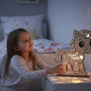 K&ouml;nnte beinhalten: Ein h&ouml;lzernes pferdef&ouml;rmiges Nachtlicht mit den Worten "Gute Nacht" und einem Herz. Das Licht projiziert Sterne und Monde an die Wand. Das Pferd steht auf einem wei&szlig;en Nachttisch.